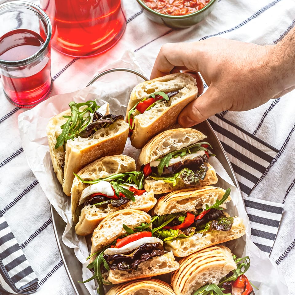 A hand reaching for a deli sandwich filled with roasted vegetables, cheese, and arugula.