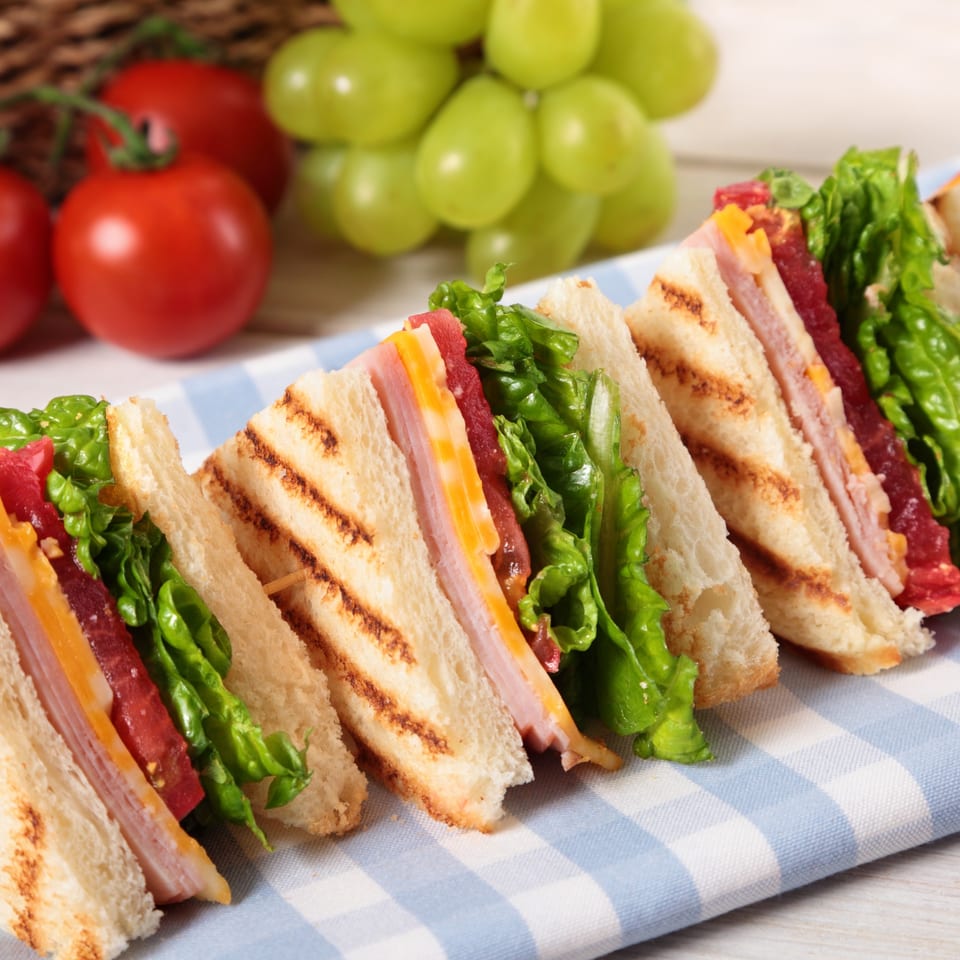 Toasted deli sandwiches with ham, cheese, lettuce, and tomato, alongside grapes and cherry tomatoes.