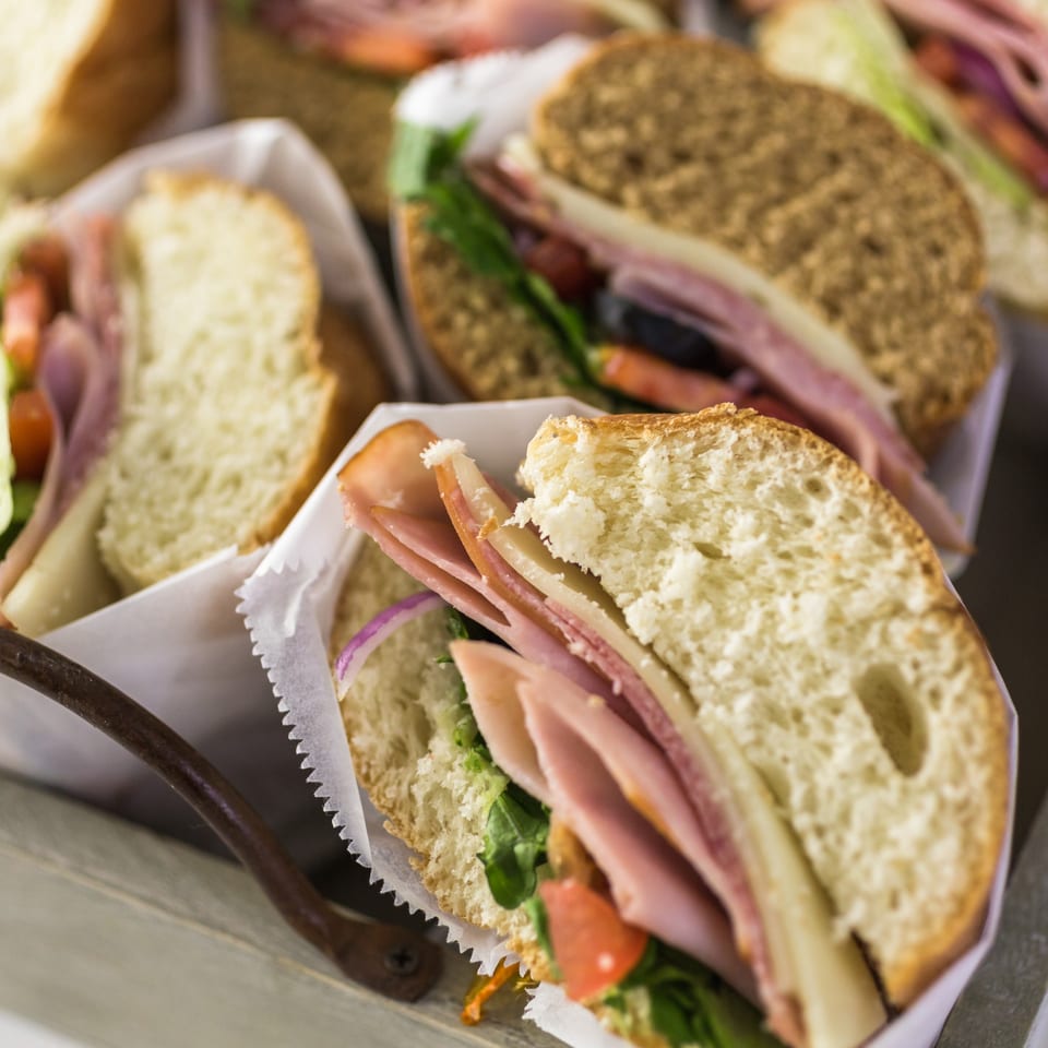 Close-up of several deli sandwiches with ham, cheese, lettuce, and tomato, wrapped in white paper.