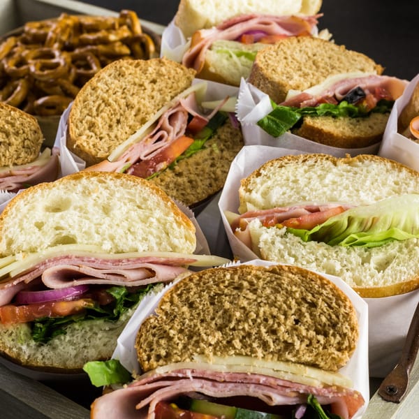 Assortment of deli sandwiches with various breads, meats, cheeses, and vegetables, alongside pretzels.