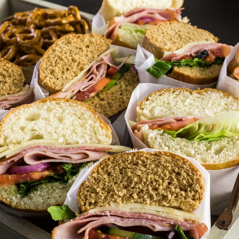 Assortment of deli sandwiches with various breads, meats, cheeses, and vegetables, alongside pretzels.